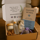 Gift box with self-care items including a candle, bottle, and card on a wooden surface.